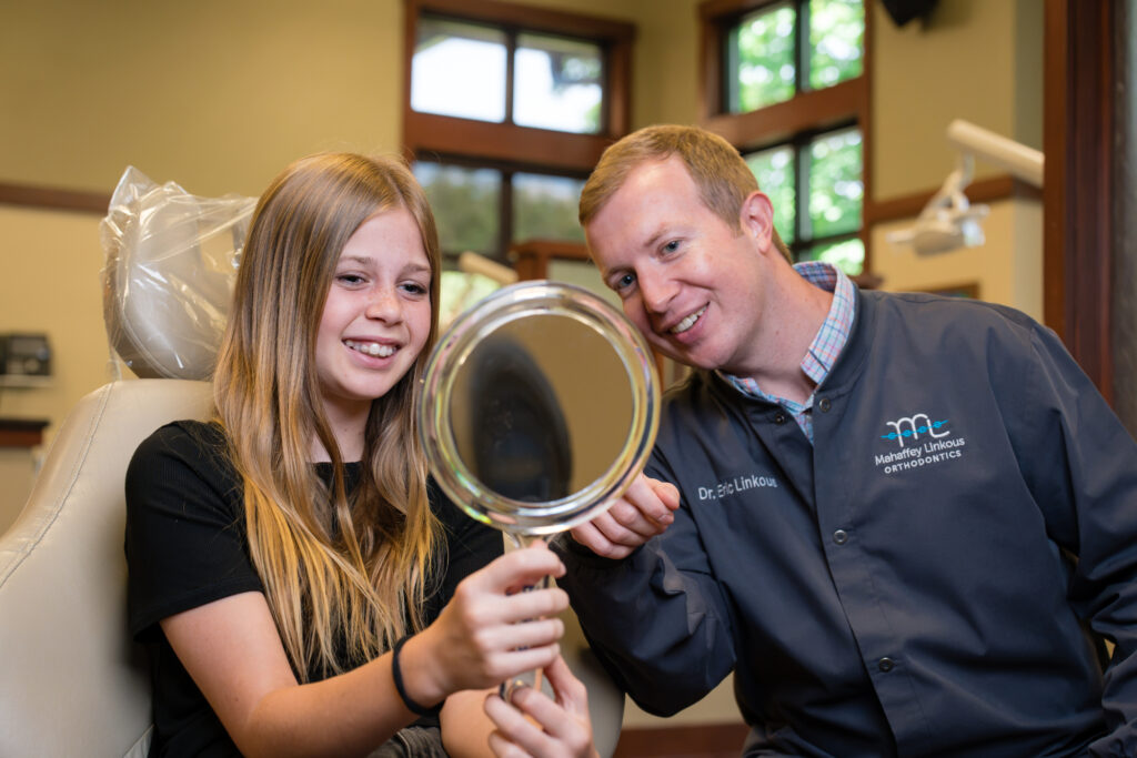 Dr. Eric Linkous smiles beside a teen patient as she checks her smile in a hand mirror during her clear aligner visit at Mahaffey Linkous Orthodontics in Peachtree City, GA.