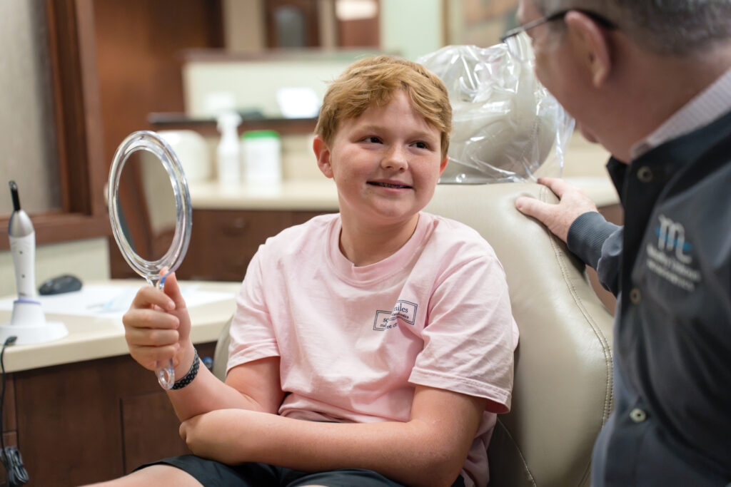 Dr. Michael Mahaffey talks with a smiling young patient in a pink shirt who holds a hand mirror in the treatment chair during a braces appointment at Mahaffey Linkous Orthodontics in Peachtree City, GA.